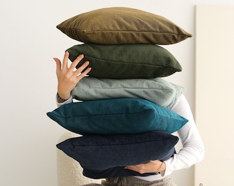 May include: A stack of five decorative throw pillows in various colors, including olive green, dark green, light blue, teal, and navy blue. The pillows are made of a soft, textured fabric.