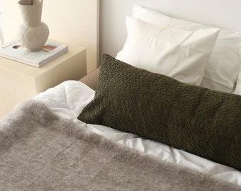 May include: A white bed with a green textured lumbar pillow and a white pillow. A gray throw blanket is on the bed. A vase with yellow dried flowers is on a wooden nightstand.