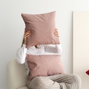 Puede incluir: Una persona sentada en un sillón blanco con textura esponjosa, sosteniendo una gran almohada rosa frente a su cara. Hay una almohada rosa más pequeña en su regazo. La persona lleva una camisa blanca de manga larga y pantalones de punto grises.