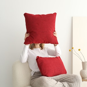 May include: A woman is sitting in a white armchair with two red throw pillows. The woman is holding a large red pillow in front of her face.