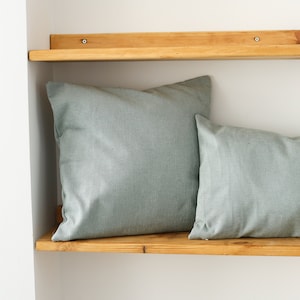 May include: Two light green throw pillows on a wooden shelf against a white wall.