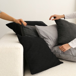 May include: A white sofa with a stack of black and grey throw cushions. The cushions are arranged in a casual, layered style.