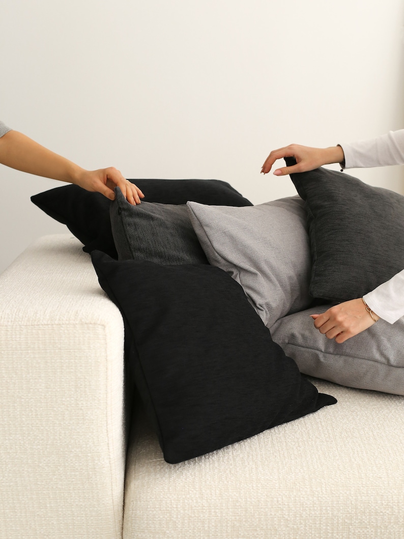 May include: A white sofa with a stack of black and grey throw cushions. The cushions are arranged in a casual, layered style.