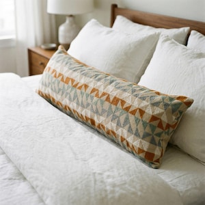 Könnte beinhalten: Ein langes, rechteckiges Zierkissen mit einem geometrischen Muster aus Dreiecken in Beige, Orange und Blau. Das Kissen liegt auf einem Bett mit weißer Bettwäsche und schafft eine gemütliche und einladende Atmosphäre. Das Kissen ist etwa 100 cm lang.