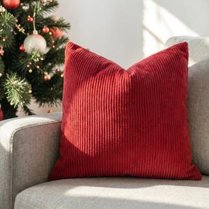 May include: A red corduroy pillow with vertical ribbing sits on a gray sofa. The pillow is square and has a soft, textured appearance. A Christmas tree with ornaments is visible in the background, suggesting a holiday theme.