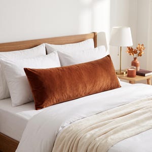 May include: A long, rectangular lumbar pillow made of rust-colored velvet fabric. The pillow is positioned horizontally across the center of a bed, resting in front of two white pillows and a white duvet cover on a wooden bed frame.