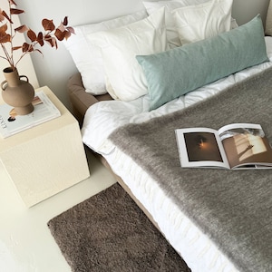 May include: A bed with white bedding and a grey throw blanket. The bed has a light green bolster pillow and two white pillows. A brown rug is on the floor in front of the bed. A small white bedside table with a magazine on top is next to the bed.