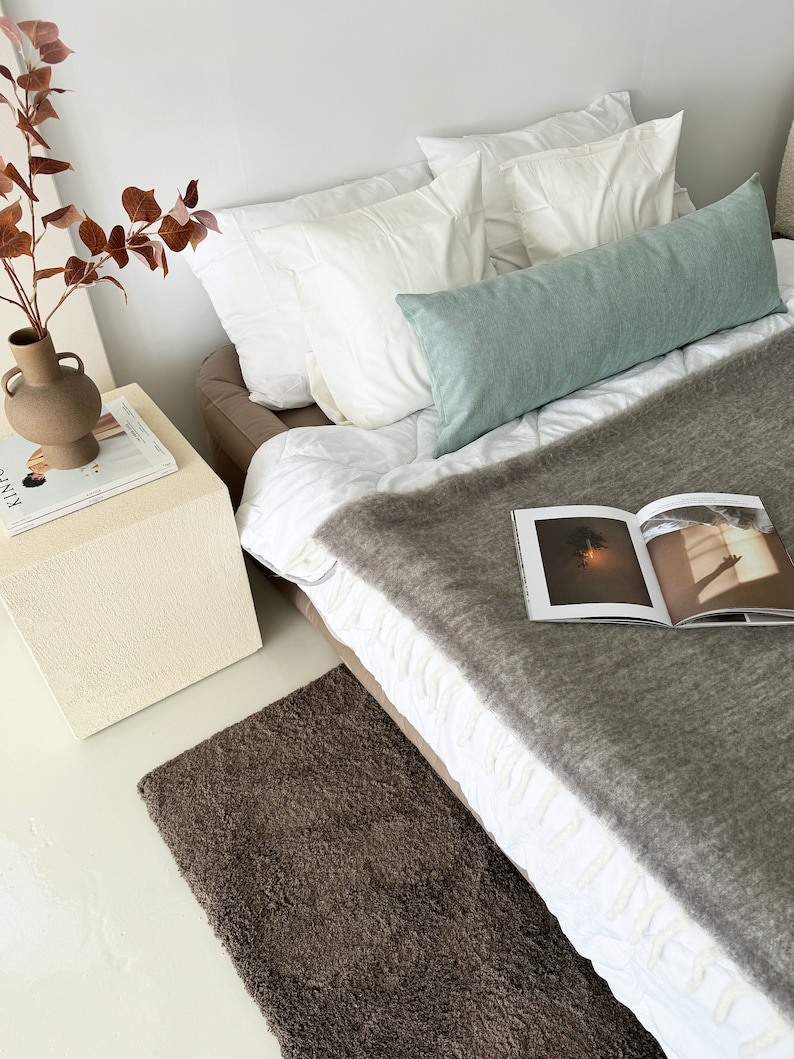 May include: A bed with white bedding and a grey throw blanket. The bed has a light green bolster pillow and two white pillows. A brown rug is on the floor in front of the bed. A small white bedside table with a magazine on top is next to the bed.