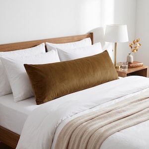 May include: A long, rectangular velvet pillow in a deep mustard brown color, positioned horizontally across a bed with white bedding, white pillows, and a light beige throw blanket, set against a wooden headboard.