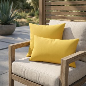 May include: Two yellow rectangular outdoor cushions sit on a beige chair cushion. The chair has a light brown wooden frame and is on a patio. A third cushion is partially visible behind the yellow cushions.