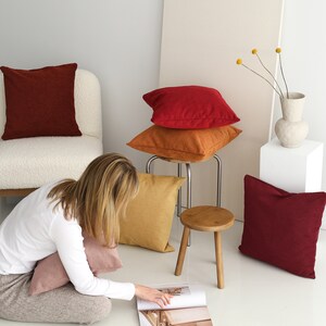 Pu&ograve; includere: Una donna &egrave; seduta sul pavimento con una rivista aperta davanti a lei. &Egrave; circondata da una variet&agrave; di cuscini decorativi in ​​toni di rosso, arancione e rosa. I cuscini sono disposti su una sedia bianca e uno sgabello di legno.