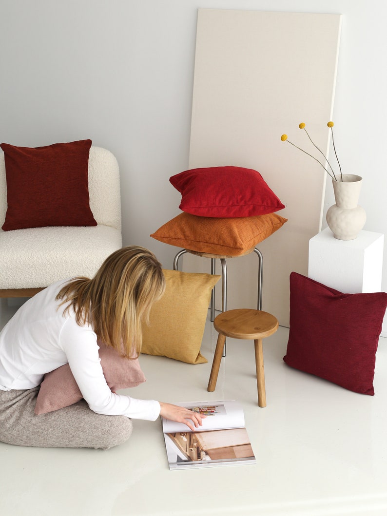 Pu&ograve; includere: Una donna &egrave; seduta sul pavimento con una rivista aperta davanti a lei. &Egrave; circondata da una variet&agrave; di cuscini decorativi in ​​toni di rosso, arancione e rosa. I cuscini sono disposti su una sedia bianca e uno sgabello di legno.