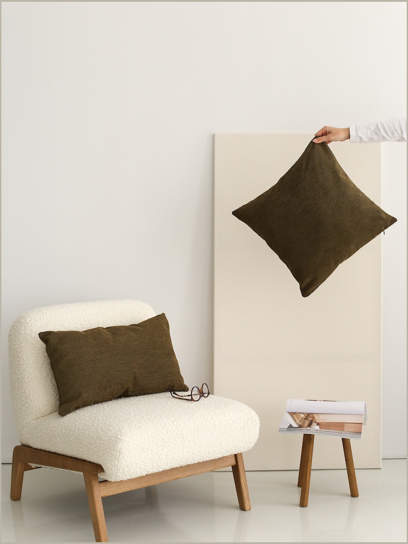 May include: A white armchair with a brown throw pillow and a wooden frame. A small wooden stool with a magazine on top sits next to the chair. A brown throw pillow is being held in the air.