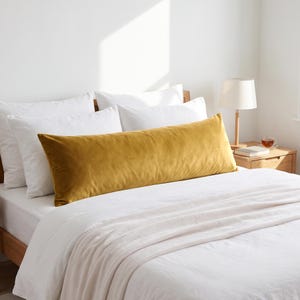 May include: A long, rectangular mustard yellow velvet body pillow positioned horizontally across a bed. The pillow features a smooth, soft-textured fabric surface and is placed in front of several white pillows on a white bed sheet and blanket.