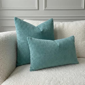 May include: Two teal corduroy cushions, one rectangular and one square, sit on a white, textured sofa. The cushions have a ribbed texture and a solid colour. The background features a white wall with moulding.