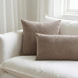 May include: Two corduroy throw pillows in a neutral taupe color rest on a white sofa. The pillows have a vertical ribbed texture. One pillow is square, and the other is rectangular. The sofa is covered in white fabric.