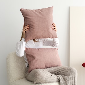 May include: A person sitting in a white fluffy armchair with two pink throw pillows. The person is holding a larger pink pillow in front of their face.