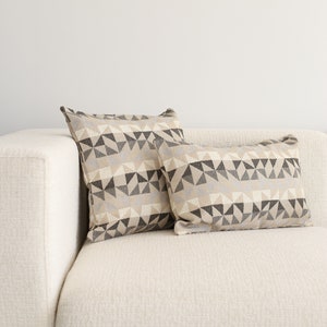 May include: Two decorative throw pillows with a geometric pattern in shades of beige, gray, and black. The pillows are on a white couch.
