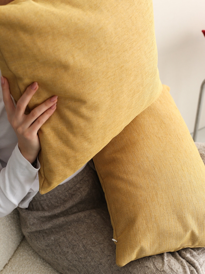 May include: Two mustard yellow throw pillows with a textured fabric. The pillows are on a brown sofa.