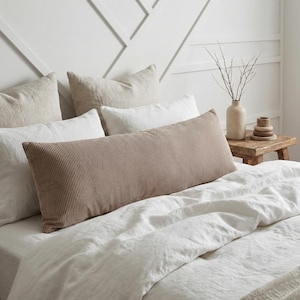 May include: A bed with white linen bedding and several pillows. A long, rectangular, corduroy pillow in a light brown color is the focal point. Other pillows are white and beige. A small wooden table holds a vase and decorative items.