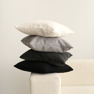 Pu&ograve; includere: Quattro cuscini decorativi impilati su un divano bianco. I cuscini sono in tonalit&agrave; di bianco, grigio e nero.