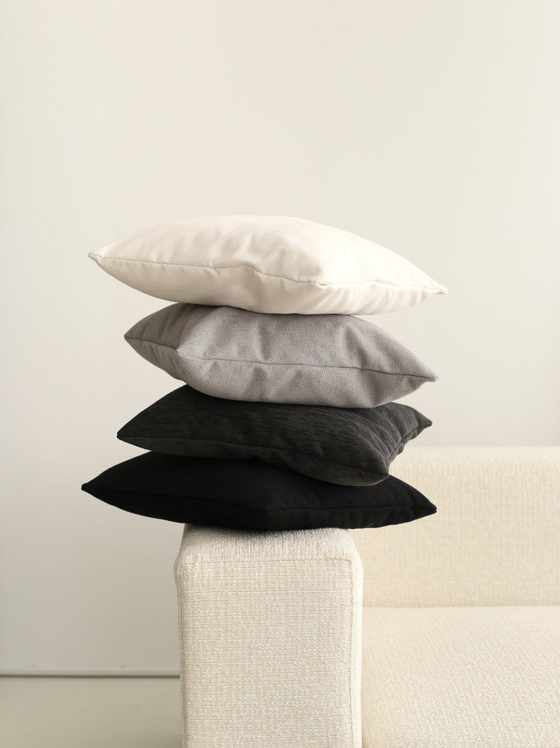 Pu&ograve; includere: Quattro cuscini decorativi impilati su un divano bianco. I cuscini sono in tonalit&agrave; di bianco, grigio e nero.