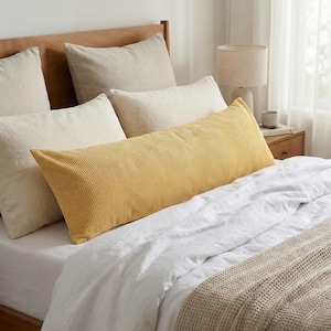 May include: A bed with a wooden headboard and several pillows. A long, mustard-yellow corduroy body pillow is centered on the bed, surrounded by white and beige pillows. White sheets and a beige blanket complete the bedding.