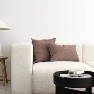May include: A white sectional sofa with two brown throw cushions. The sofa is in a modern living room with a black side table and a brown rug.