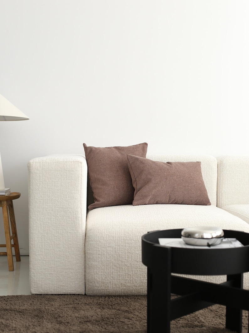 May include: A white sectional sofa with two brown throw cushions. The sofa is in a modern living room with a black side table and a brown rug.