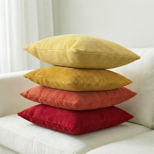 May include: Four corduroy cushions stacked on a white sofa. The cushions are in shades of yellow, orange, and red. The top cushion is light yellow, the second is a darker yellow, the third is orange, and the bottom is red.