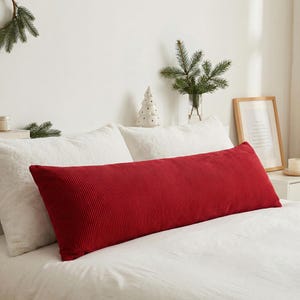 Può includere: Un lungo cuscino rettangolare di velluto a coste rosso è appoggiato su un letto bianco. Il cuscino è il punto focale, con una superficie strutturata e una tonalità rosso intenso. Il letto è fatto con lenzuola bianche e lo sfondo include elementi decorativi.