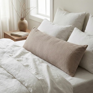 May include: A long, rectangular, corduroy pillow in a neutral beige color rests on a bed with white linens and pillows. The pillow has a ribbed texture and is positioned horizontally. The scene suggests a bedroom setting with natural light.