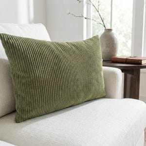 Puede incluir: Un cojín rectangular de pana verde oliva descansa sobre un sillón blanco. El cojín tiene una textura acanalada y está colocado contra el respaldo de la silla. Un jarrón y un libro son visibles en el fondo.