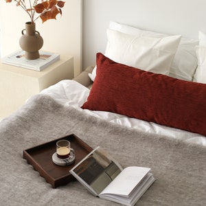 Pu&ograve; includere: Una coperta di lana grigia su un letto con un cuscino rosso, un vassoio di legno con un bicchiere di caff&egrave; e un libro.
