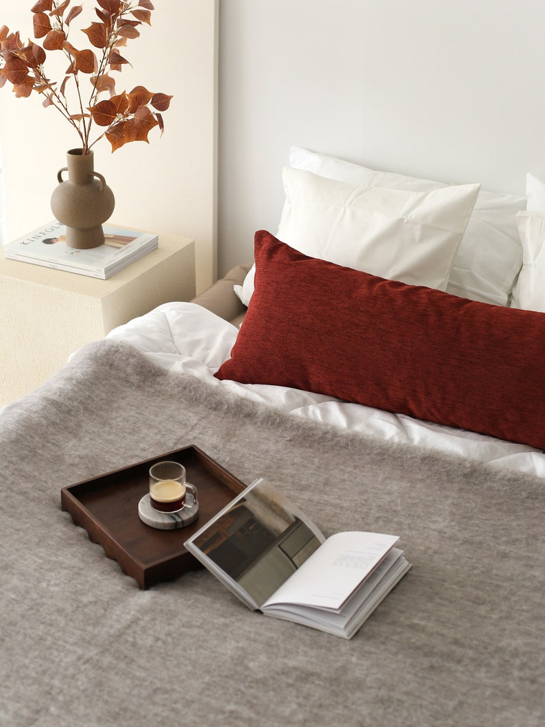 Pu&ograve; includere: Una coperta di lana grigia su un letto con un cuscino rosso, un vassoio di legno con un bicchiere di caff&egrave; e un libro.