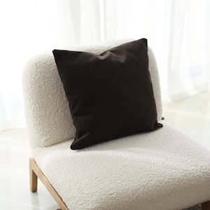 May include: A white fluffy armchair with a brown throw pillow. The chair has wooden legs and is sitting on a brown rug.