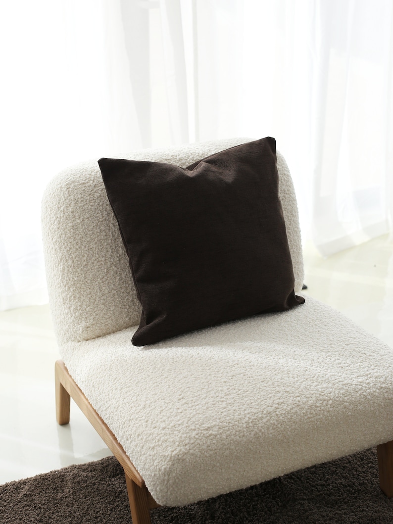 May include: A white fluffy armchair with a brown throw pillow. The chair has wooden legs and is sitting on a brown rug.