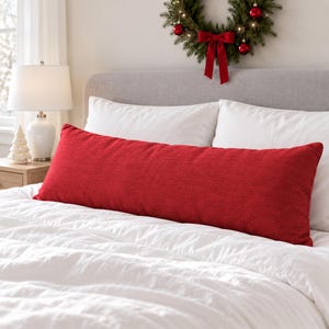 May include: A long, rectangular lumbar pillow in a solid, deep red textured fabric. The pillow is positioned horizontally across the center of a bed, resting in front of two white pillows on a white duvet cover.