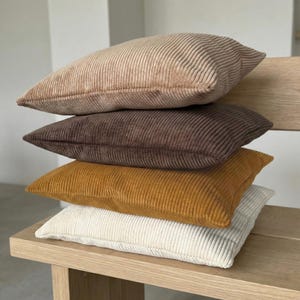 May include: A stack of four corduroy pillows in varying earth tones: beige, brown, mustard yellow, and off-white. The pillows are rectangular and have a ribbed texture. They are stacked on a light-colored wooden bench.