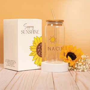 May include: A clear glass water bottle with a bamboo lid and straw. The bottle has a sunflower graphic and the text "Sipping Sunshine in a Cup". The bottle is sitting on a white coaster. The bottle is packaged in a white box with a sunflower graphic and the text "Sipping Sunshine".