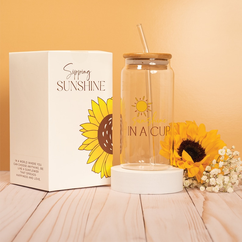 May include: A clear glass water bottle with a bamboo lid and straw. The bottle has a sunflower graphic and the text "Sipping Sunshine in a Cup". The bottle is sitting on a white coaster. The bottle is packaged in a white box with a sunflower graphic and the text "Sipping Sunshine".