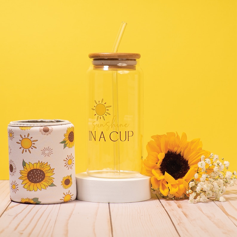May include: A clear glass tumbler with a bamboo lid and straw. The tumbler has a sunflower and the text "Sunshine in a Cup" printed on it. The tumbler is sitting on a white coaster. There is a sunflower and white flowers in the background.