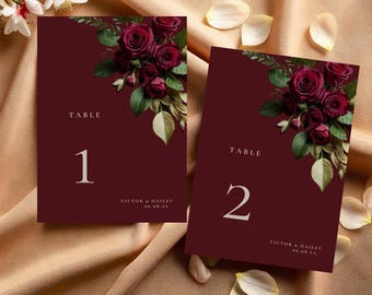 May include: Burgundy table number cards with floral arrangements of dark red roses and green leaves. The cards have the numbers 1 and 2 in white, with the names "Victor & Hailey" and a date at the bottom. The cards are on a gold satin surface.