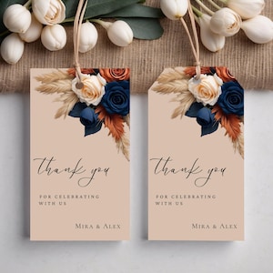 May include: Two beige thank you tags with a floral design. The flowers are white, orange, and blue. The text on the tags reads "Thank you for celebrating with us, Mira & Alex".