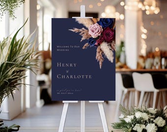 May include: A navy blue wedding welcome sign on a white easel. The sign features the text "Welcome to Our Wedding" and the names "Henry & Charlotte" with the date. The sign is decorated with a floral arrangement of blue, pink, and burgundy flowers.