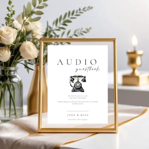 May include: A white sign with black text that reads "AUDIO guestbook" and features a black rotary phone illustration. The sign is in a gold frame and is sitting on a table with white flowers.