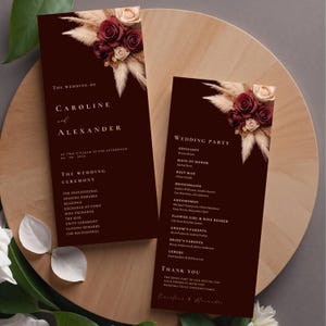 Peut inclure: Programme de cérémonie de mariage bordeaux avec un motif floral composé de pampas séchées et de roses bordeaux. Le programme liste la fête de mariage et l'ordre de la cérémonie.