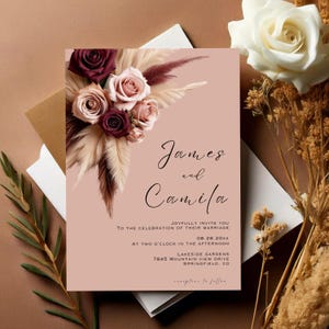 May include: A blush pink wedding invitation with a floral design featuring burgundy and blush roses, beige feathers, and the text "James and Camila" in black calligraphy. The invitation includes the date "08.28.20XX", the time "Two o'clock in the afternoon", the location "Lakeside Gardens, 7845 Mountain View Drive, Springfield, CO", and the text "Reception to follow".