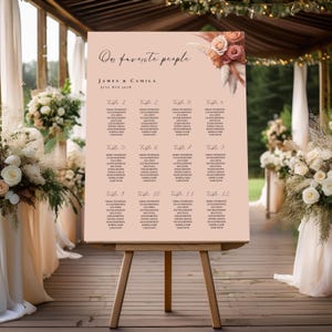 May include: A wedding seating chart on a wooden easel. The sign is a light peach color with the words "Our favorite people" in elegant script. The names James & Camila and the date are also displayed. Floral accents adorn the top right corner.
