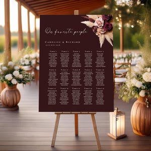 Op de afbeelding: Een bordeauxrode trouwzittingstafel op een houten ezel. De tafel bevat de tekst "Our favorite people" en de namen van het bruidspaar, Caroline & Richard, met een lijst met gastnamen per tafel. Bloemenaccenten sieren de rechterbovenhoek.